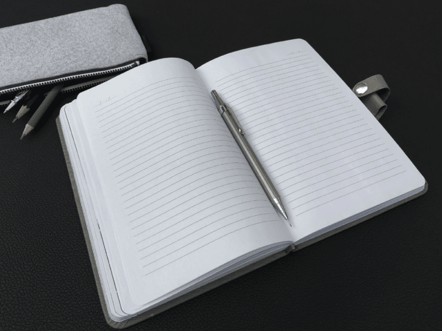 A notebook open with a sliver pen and a grey pencil case with pencils in it